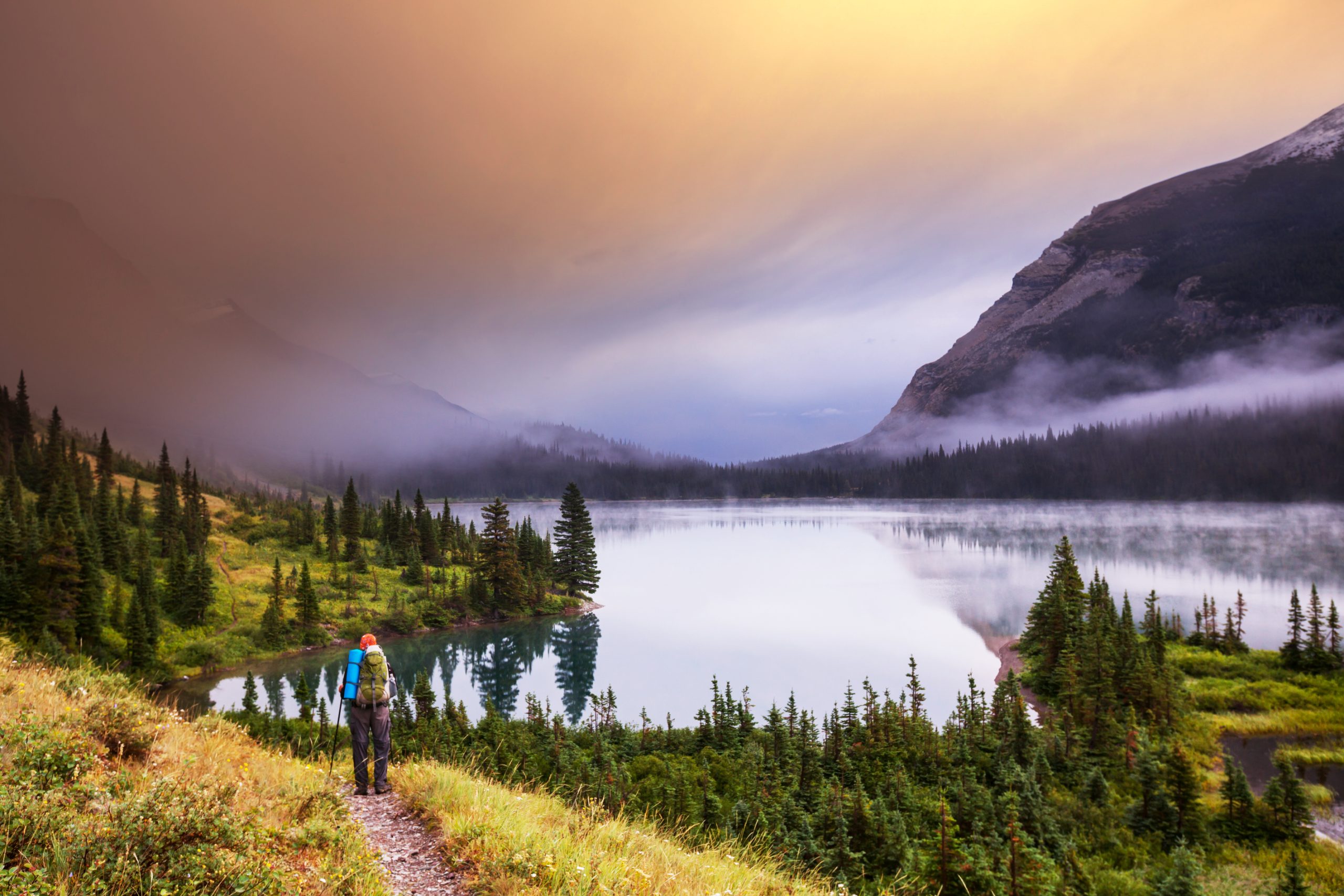 A mountain lake, with mountains in the background and a person hiking along the shore on a path.
