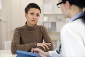 A person sits at a table with arms crossed, speaking to a healthcare professional holding a blue clipboard and pen in a medical office setting.