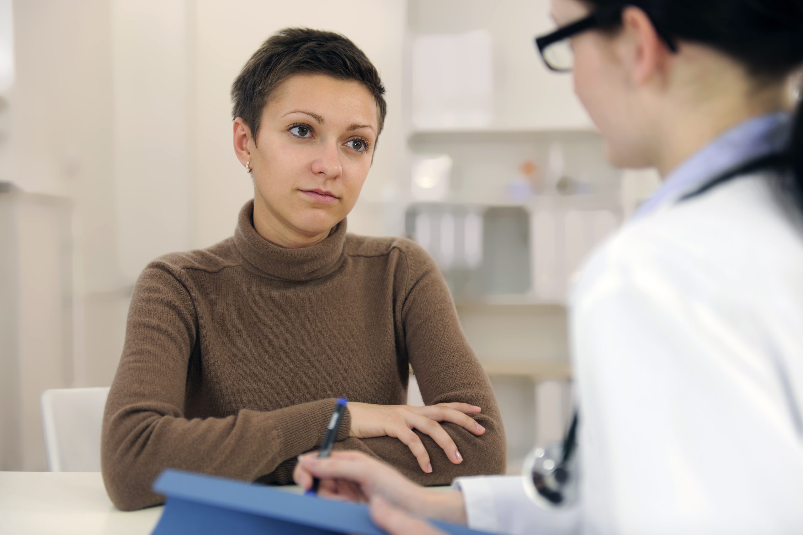 A person sits at a table with arms crossed, speaking to a healthcare professional holding a blue clipboard and pen in a medical office setting.